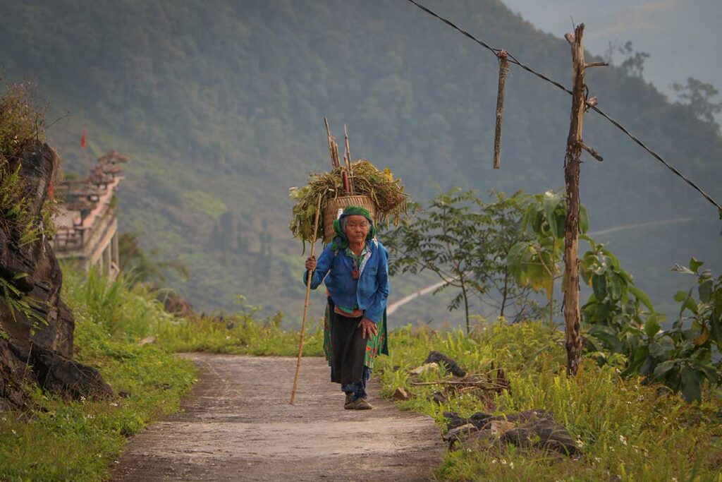 Elderly ethnic woman carrying a basket on a mountain trail near Ha Giang Aya Lodge.