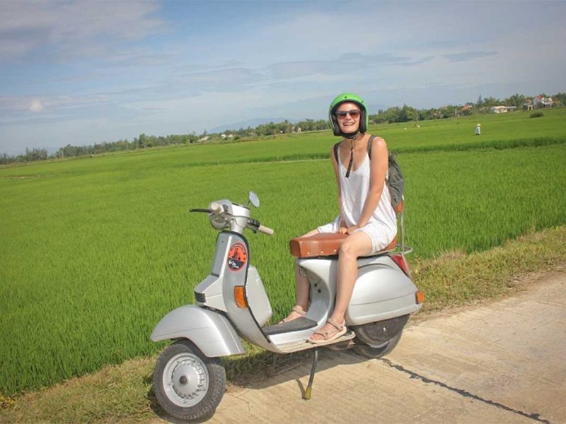 hoi an motorbike tour by vespa 1 Hoi An Vespa tour through green rice fields with a traveler riding on the back of a vintage scooter
