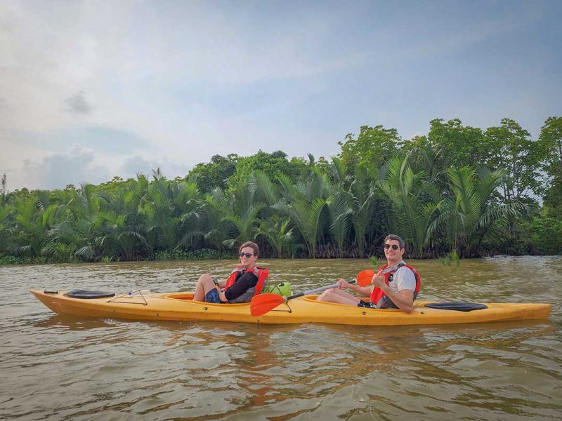 kayaking tour hoi an mangrove forest 15 Two travelers kayaking across calm brown river water with mangrove palms in the background near Hoi An