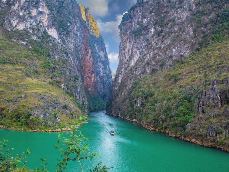 ma pi leng pass 8 A distant view of a boat gliding through the emerald-green waters of Nho Que River, surrounded by the dramatic cliffs of Ma Pi Leng Pass.