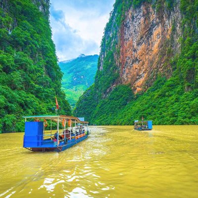 A tourist boat cruising along the brown waters of the Nho Que River, passing between the towering cliffs of Ma Pi Leng Pass in Ha Giang.