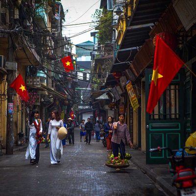 Ta Hien Street, Hanoi Old Quarter: A street vendor carries a traditional shoulder pole laden with baskets of fruit on Ta Hien Street in Hanoi's Old Quarter. Vietnamese women in white Ao Dai stroll past colorful shops adorned with Vietnamese flags.