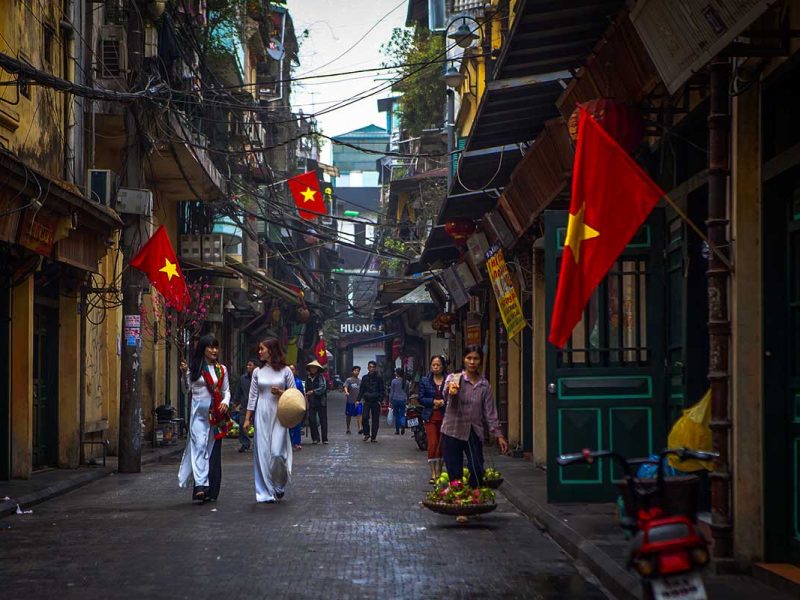 Ta Hien Street, Hanoi Old Quarter: A street vendor carries a traditional shoulder pole laden with baskets of fruit on Ta Hien Street in Hanoi's Old Quarter. Vietnamese women in white Ao Dai stroll past colorful shops adorned with Vietnamese flags.