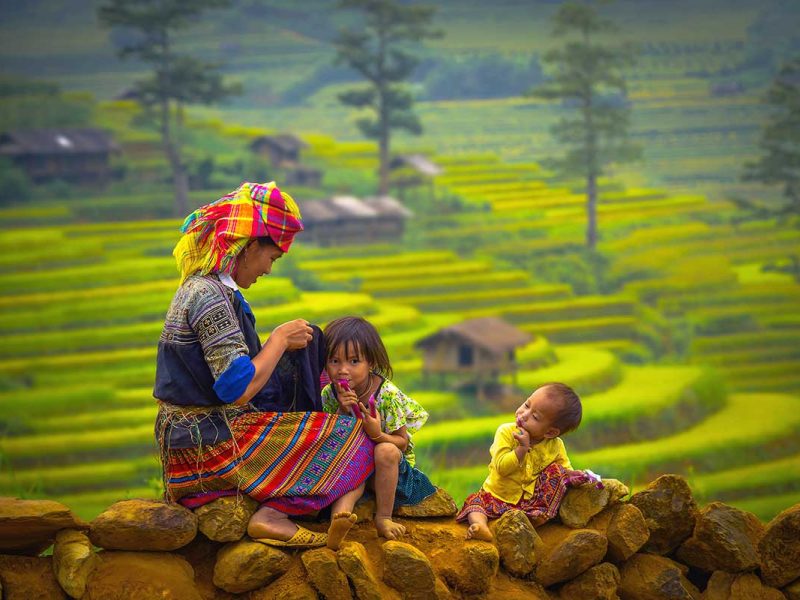 sapa minority rice fields 1 A colorful ethnic minority woman sitting on a stone wall with two children, with terraced rice fields of Sapa in the background.